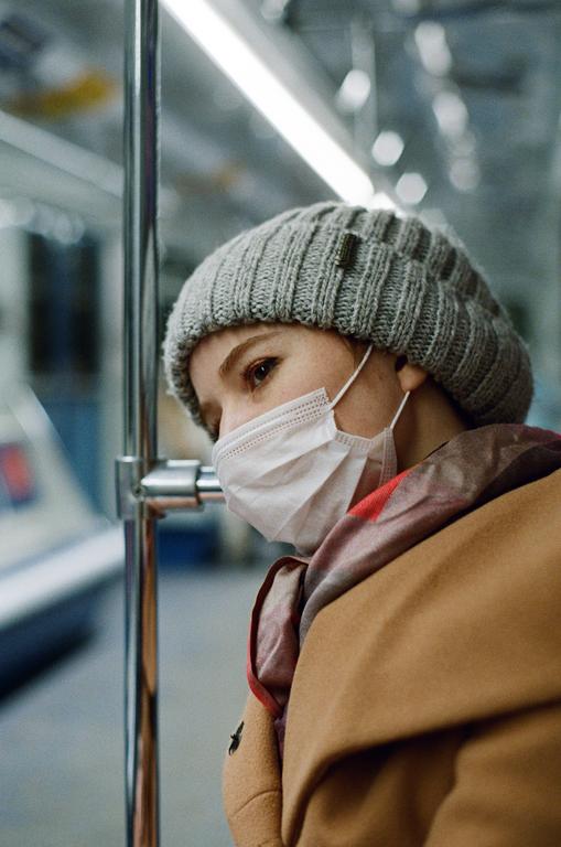 A woman wearing a face mask and a knit hat looks out a window while riding public transportation.