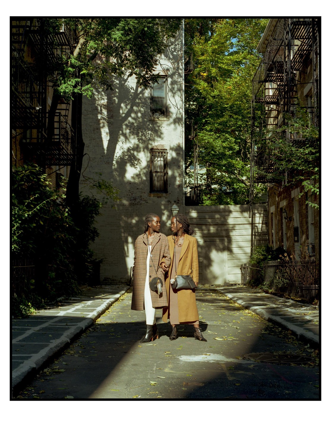 Two women stand on a street between buildings with fire escapes and trees, looking at each other.