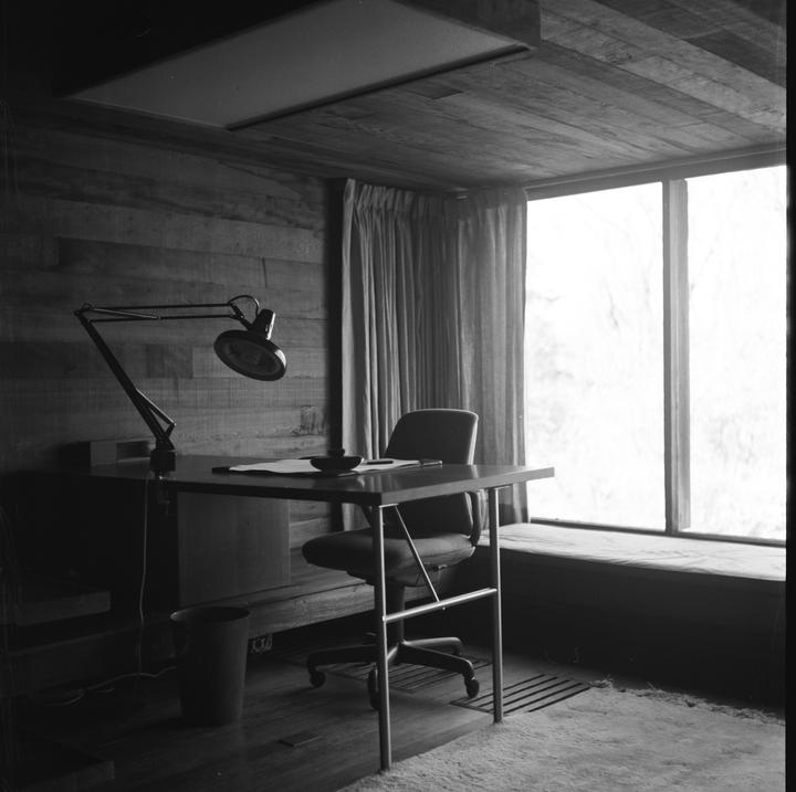A desk with a chair and a lamp is positioned in front of a large window with curtains.