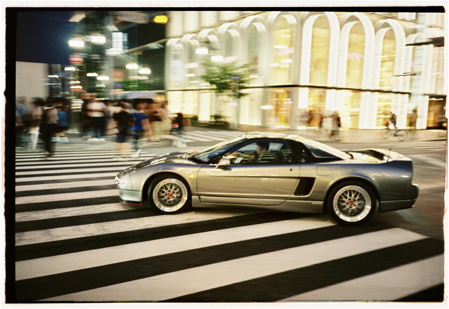 A silver sports car drives across a crosswalk at night with blurred people and buildings in the background.