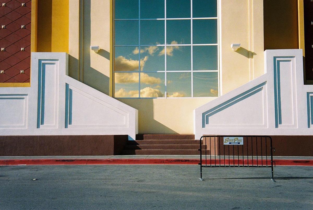 Clouds are reflected in the windows of a building with a red curb and a black barrier in the foreground.