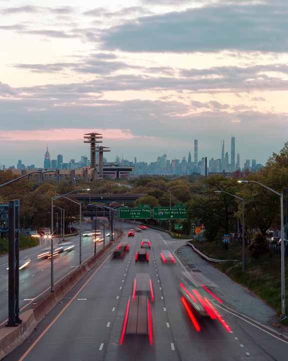 A highway with blurred car lights leads towards a city skyline with prominent towers under a cloudy sunset sky.