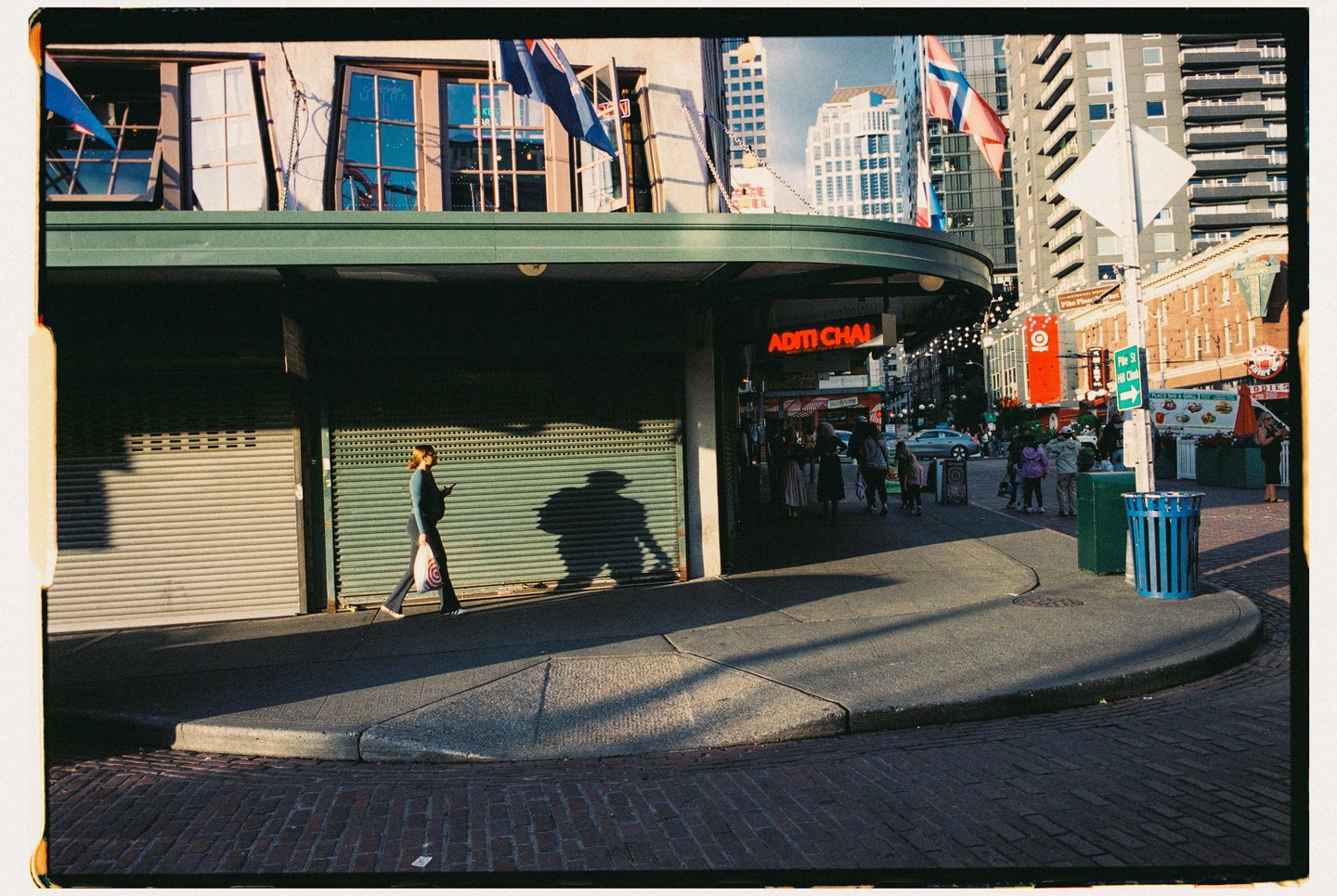A woman walks on a city sidewalk past closed storefronts with a large shadow cast beside her.