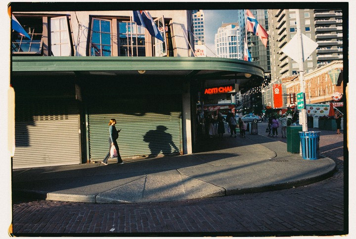 A woman walks on a city sidewalk past closed storefronts with a large shadow cast beside her.
