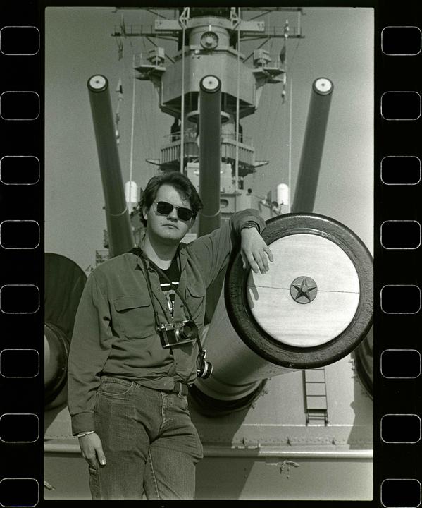 A man wearing sunglasses and a camera around his neck leans on a large gun turret on a battleship.