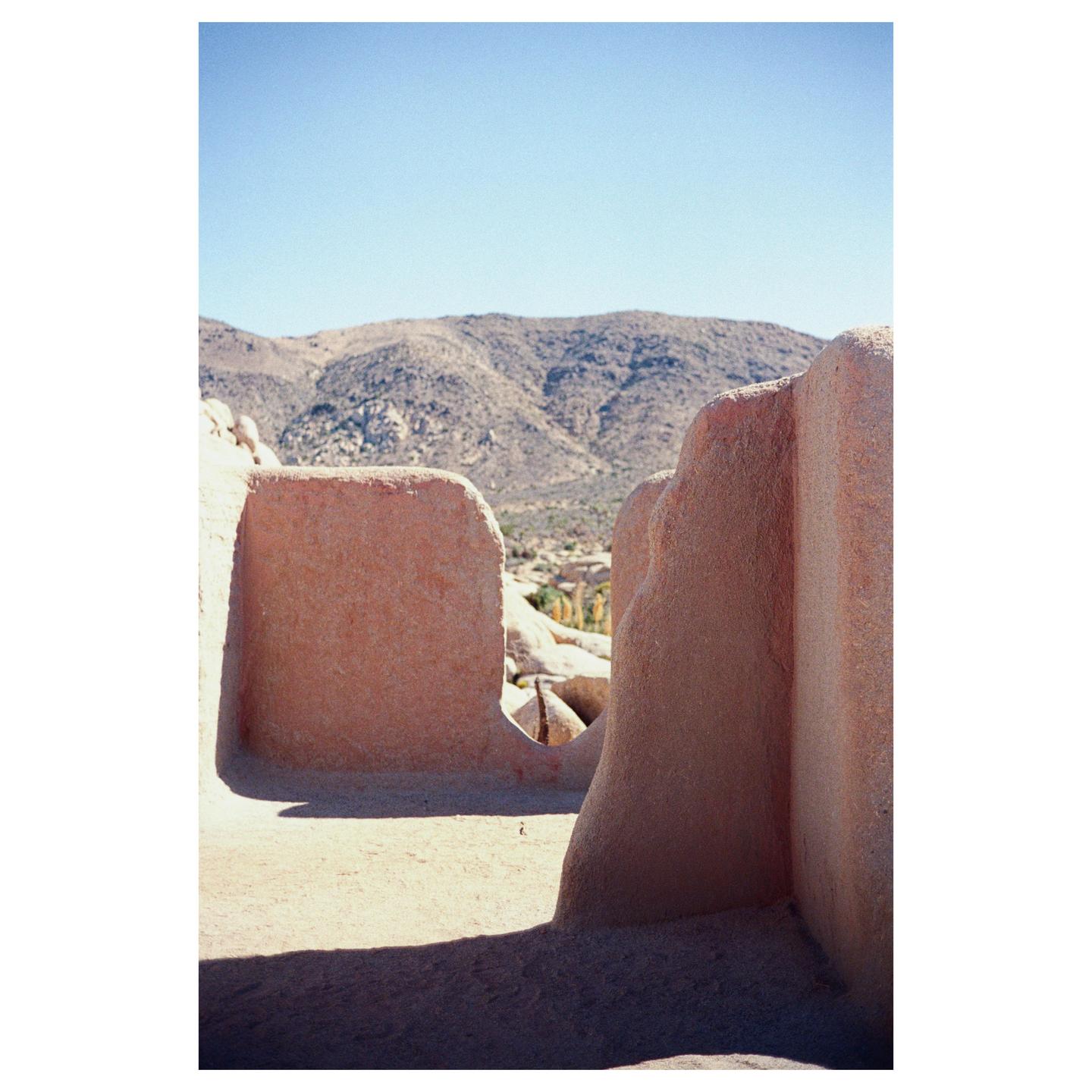 A desert landscape with mountains in the background is framed by two adobe walls in the foreground.