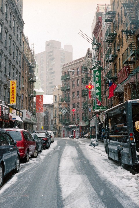 A snow-covered street in Chinatown is lined with parked cars and tall buildings with fire escapes.