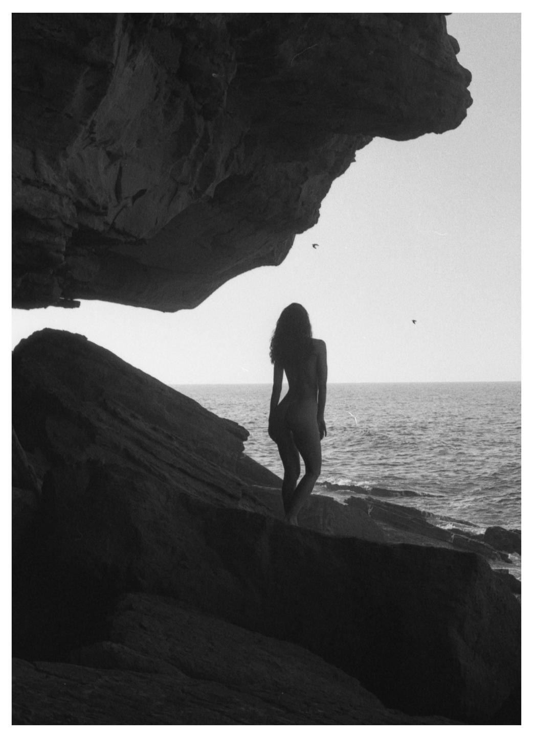 A woman stands on a rocky cliff overlooking the ocean, with a large rock formation above her.
