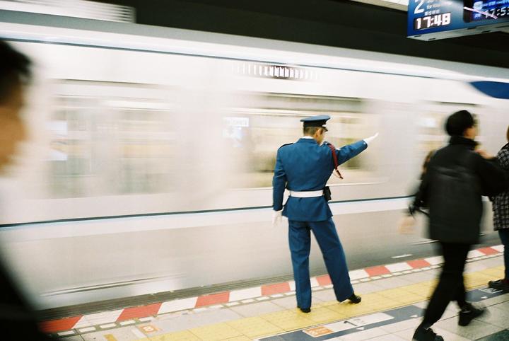 A man in a blue uniform stands on a train platform with a moving train blurring past.