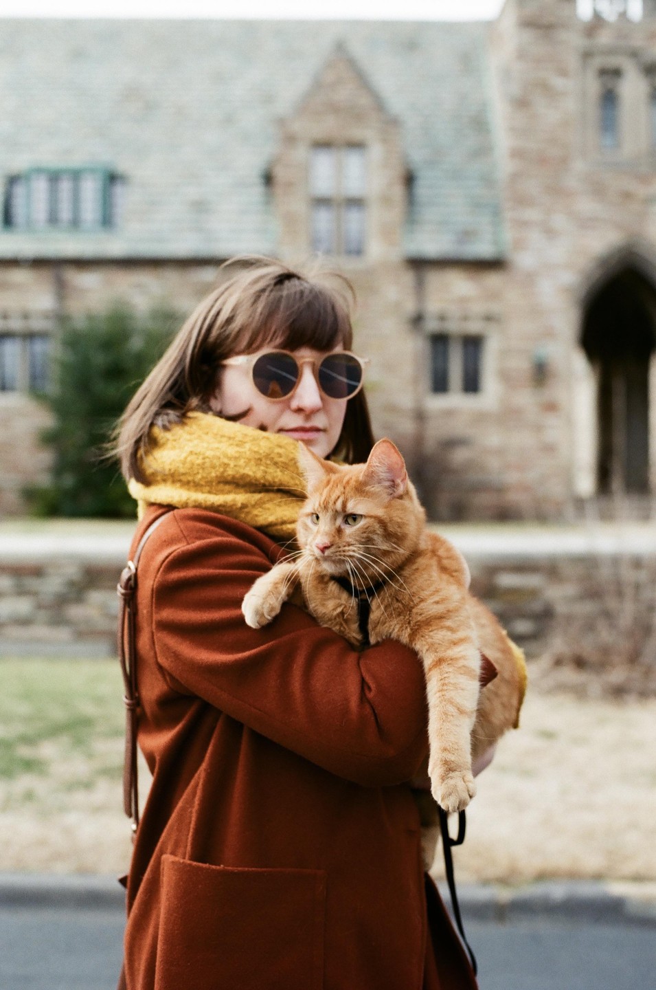 A woman wearing sunglasses and a brown coat holds an orange cat in front of a stone building.