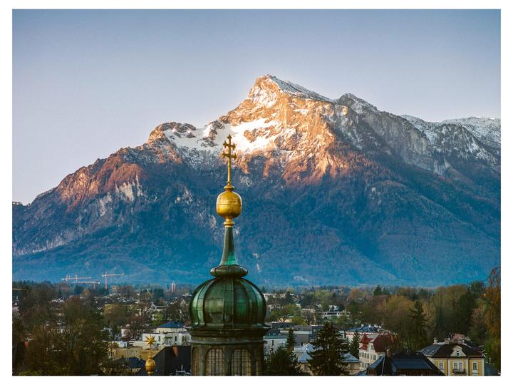 A golden church spire rises above a town with snow-capped mountains in the background.