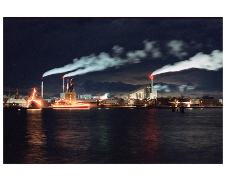 A body of water reflects the lights of a city at night, with smoke rising from industrial buildings.
