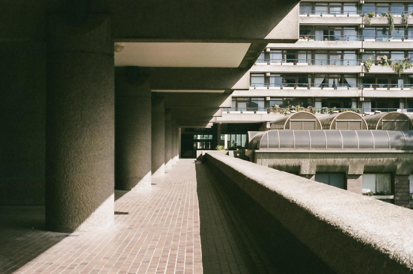 A long concrete walkway with pillars on the left and a concrete railing on the right leads to a building with many windows and balconies.