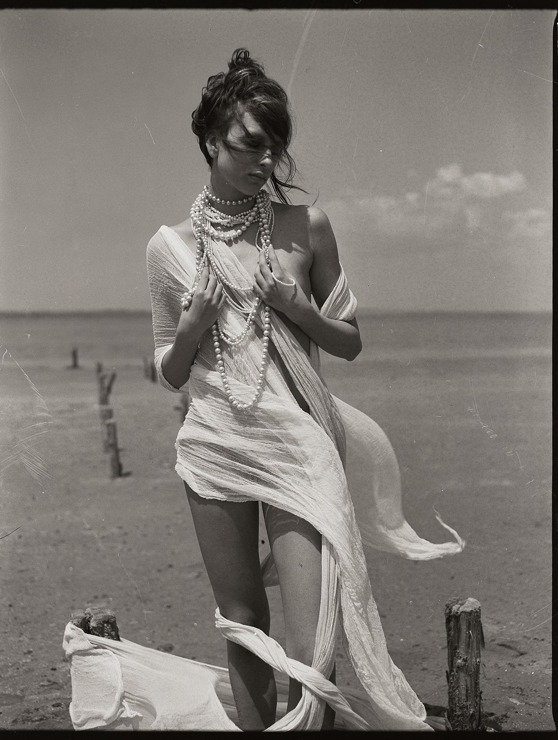 A woman wearing many pearl necklaces and draped in sheer fabric stands on a sandy shore with wooden posts in the water.