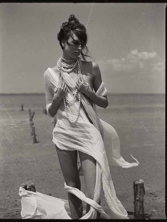 A woman wearing many pearl necklaces and draped in sheer fabric stands on a sandy shore with wooden posts in the water.