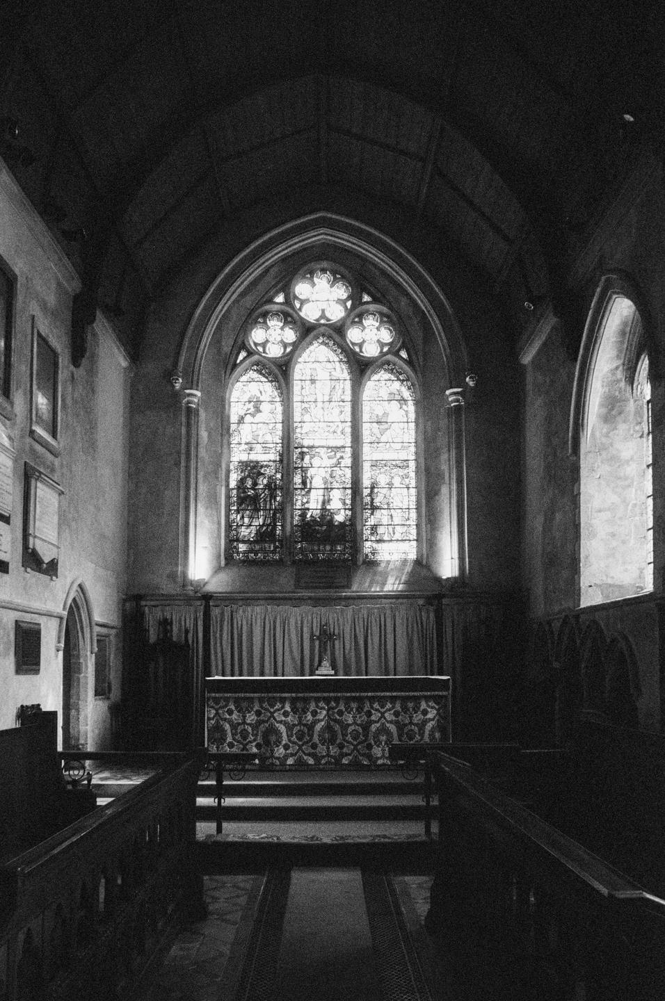 A black and white photograph shows the interior of a church with a large stained glass window at the far end.