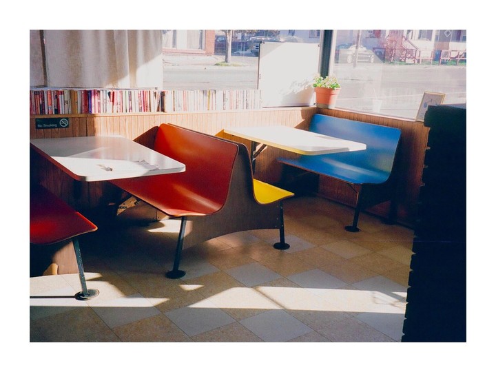 Sunlight streams into a diner with red, yellow, and blue booths and tables, with a bookshelf lining the wall.