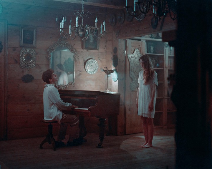 A man plays a piano while a woman stands barefoot in a wooden room with chandeliers and mirrors.