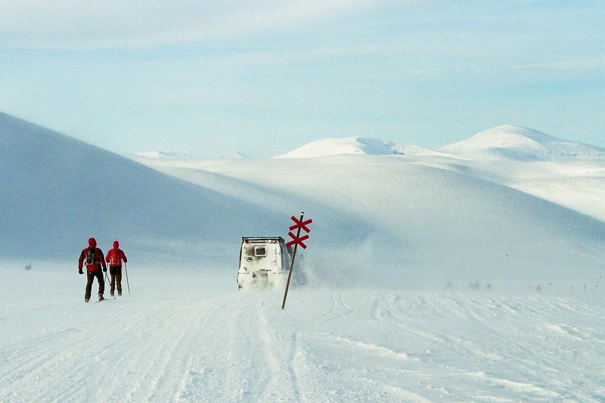 Two people on skis are next to a white vehicle on a snowy mountain landscape with a red cross sign.