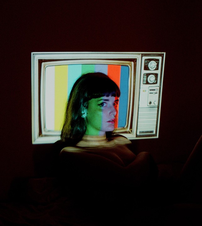 A woman with bangs looks to the side as a television test pattern is projected onto her and the wall behind her.