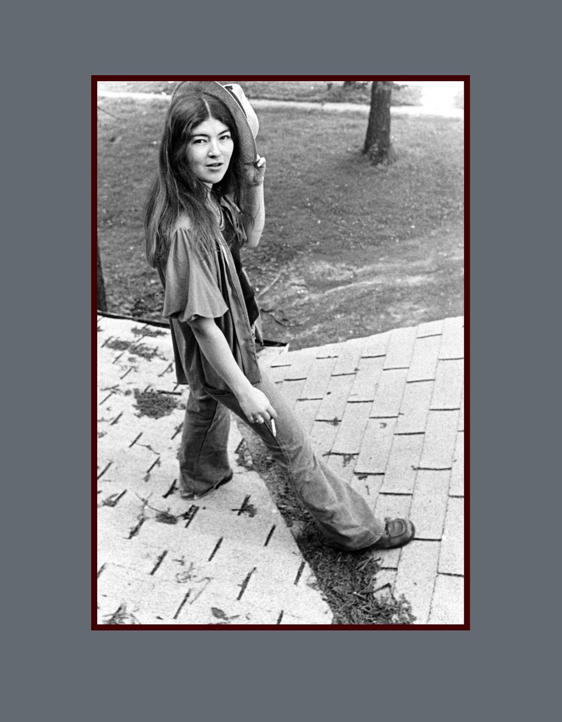 A woman stands on a shingled roof, holding a hat and looking over her shoulder.