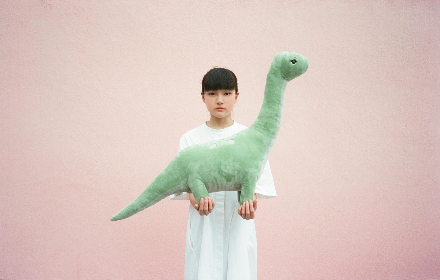 A girl in a white dress holds a large green dinosaur toy in front of a pink wall.
