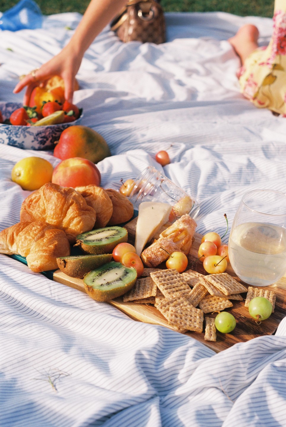 A picnic blanket is spread with croissants, fruit, crackers, cheese, and a glass of white wine.