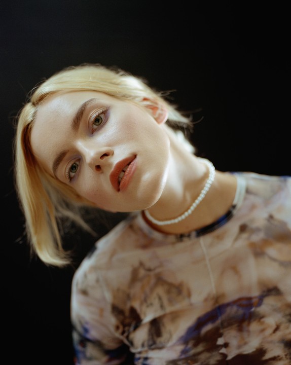 A woman with blonde hair and a pearl necklace looks directly at the viewer.