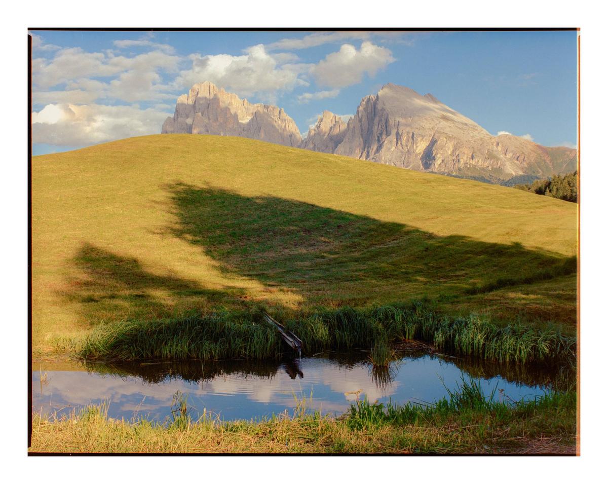 A small pond with grass around it sits in the foreground of a grassy hill with mountains in the background under a blue sky with clouds.