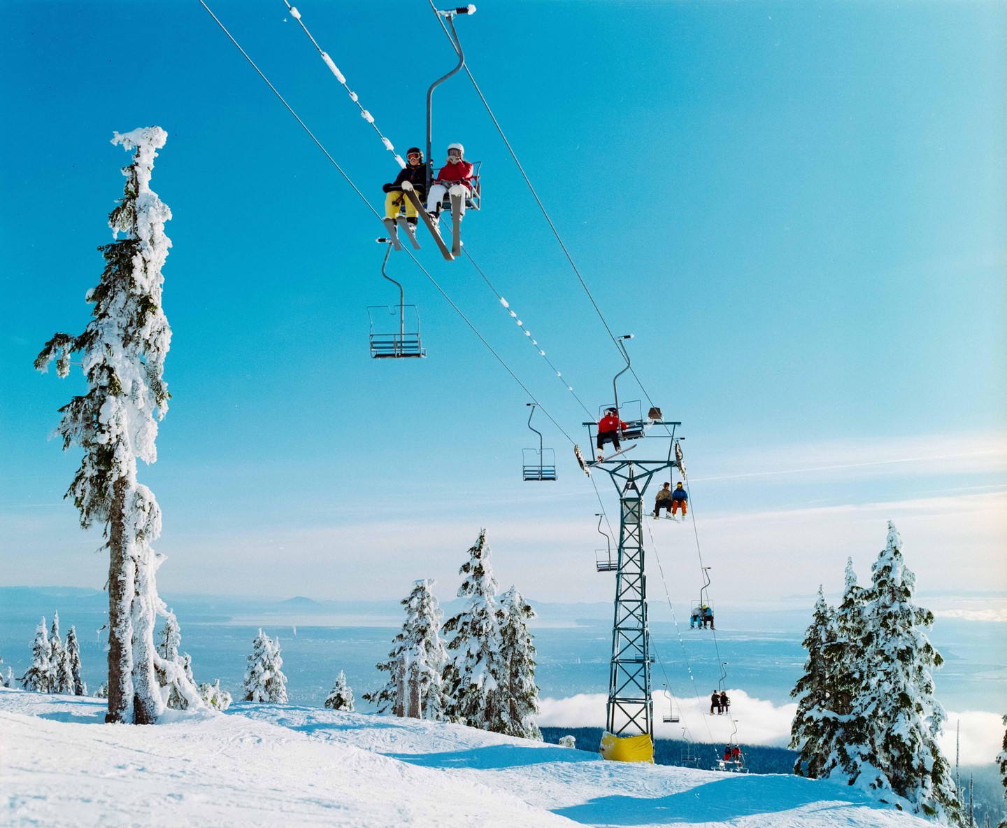People ride a ski lift up a snowy mountain with snow-covered trees and a distant body of water under a clear blue sky.