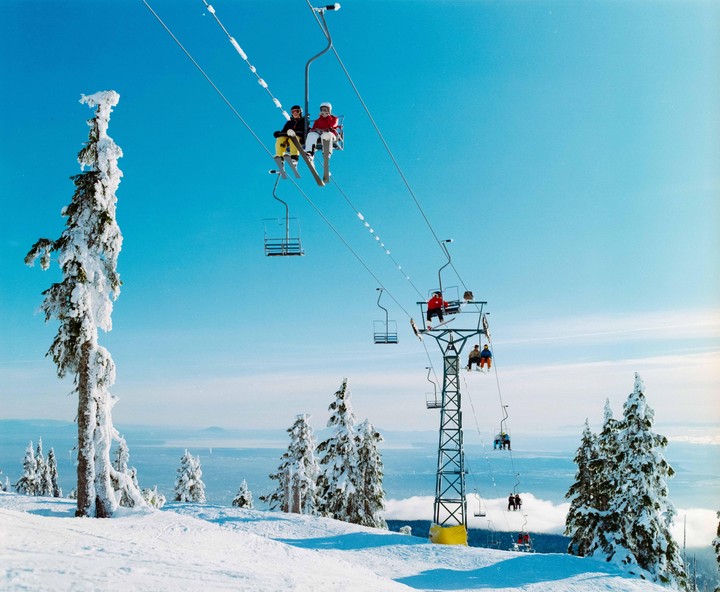 People ride a ski lift up a snowy mountain with snow-covered trees and a distant body of water under a clear blue sky.