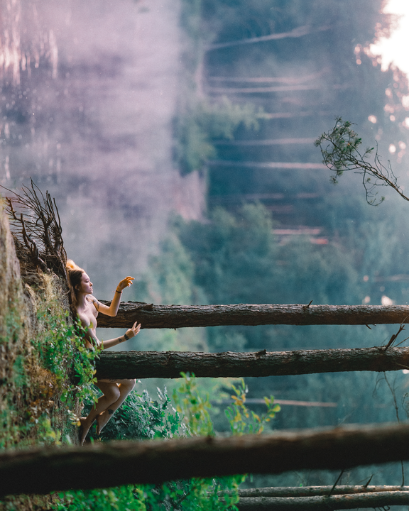 A woman with her arms outstretched sits on a cliffside with trees and fog in the background.