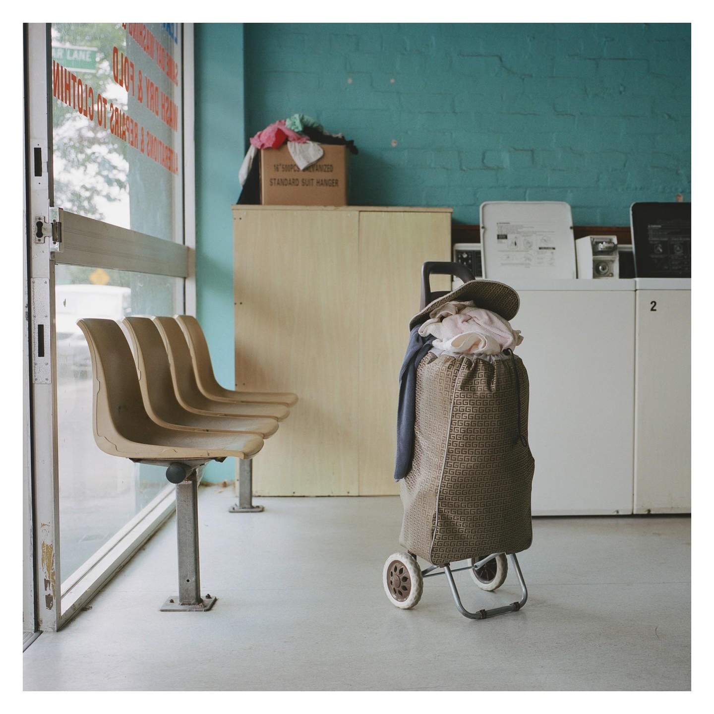 A laundry cart filled with clothes sits in a laundromat next to a row of empty seats.