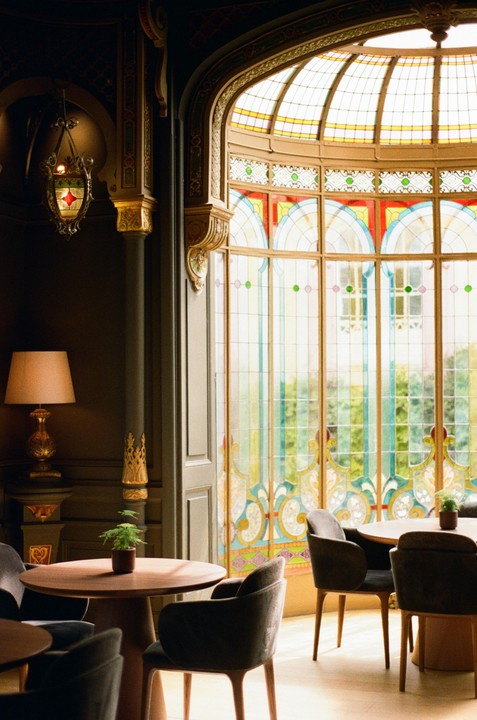 A room with ornate decor and stained glass windows features several tables and chairs.