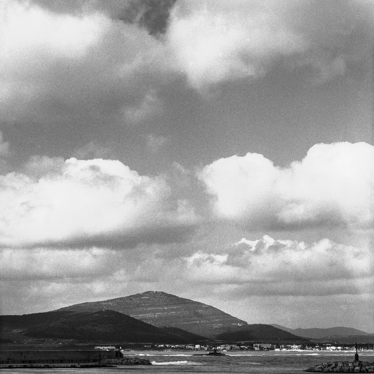Three hills rise above a body of water with a town at its base under a cloudy sky.