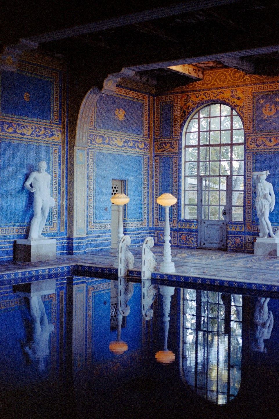 A blue tiled indoor pool with statues and reflections is illuminated by natural light from a large arched window.