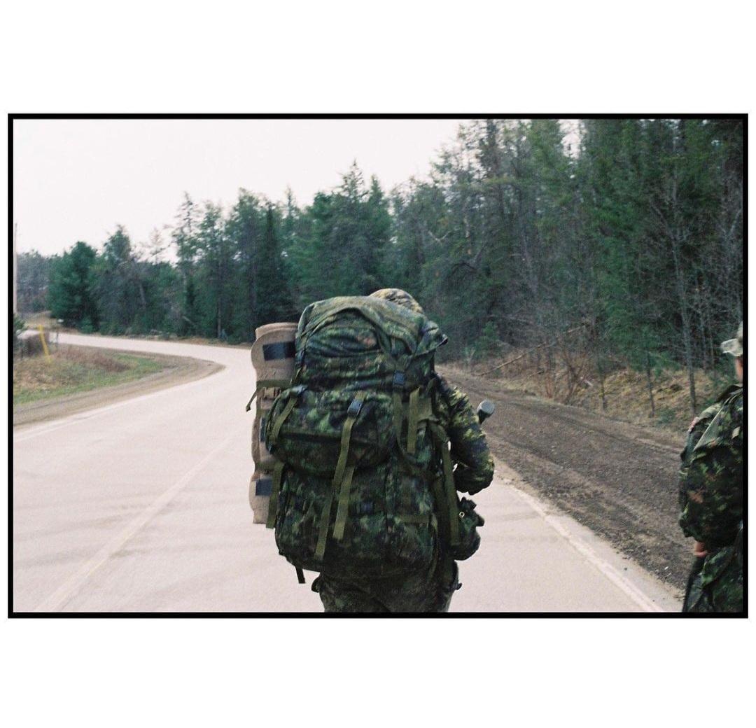 A person in camouflage with a large backpack walks along a road next to a forest.