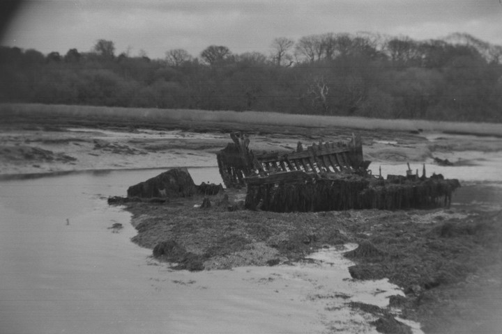 A decaying shipwreck rests on a muddy shore next to a body of water, with a distant treeline under a cloudy sky.