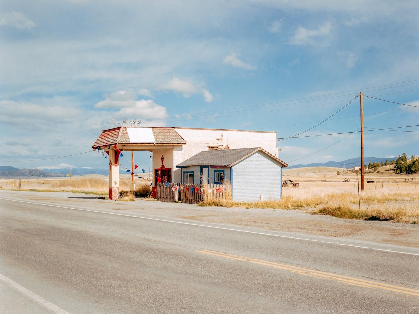 An old gas station with a small blue building next to it stands by a road in a dry, grassy landscape under a partly cloudy sky.