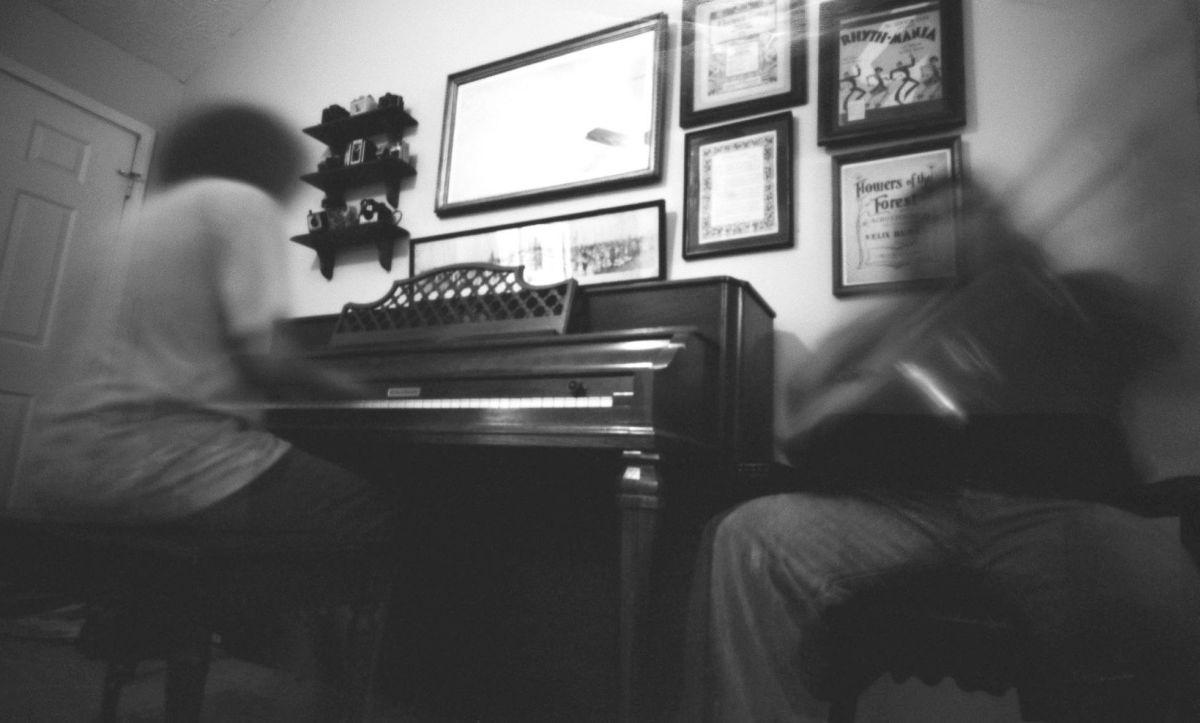A blurry image shows a person playing a piano and another person playing a guitar in a room with framed pictures on the wall.