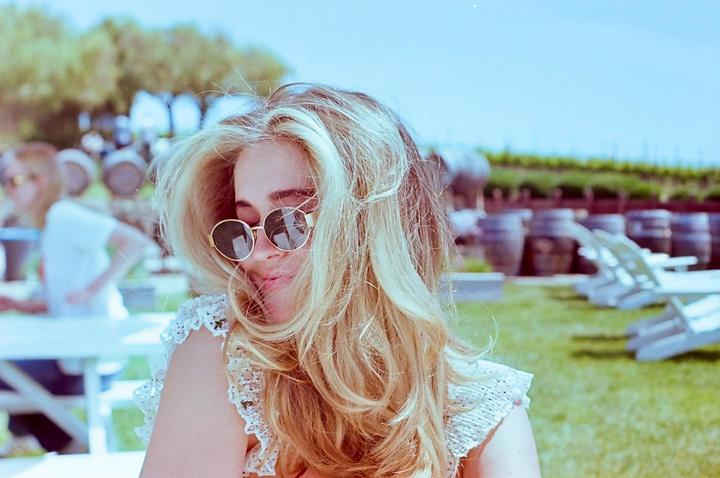 A woman with long blonde hair and sunglasses smiles while sitting outdoors with wine barrels in the background.