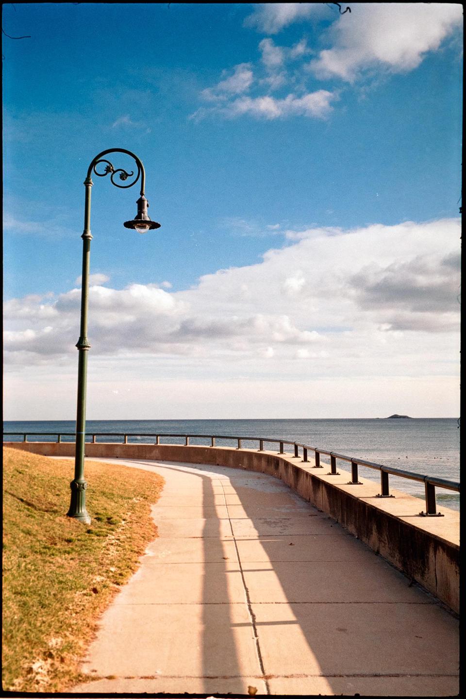 A curved path with a lamppost runs alongside a body of water under a partly cloudy sky.