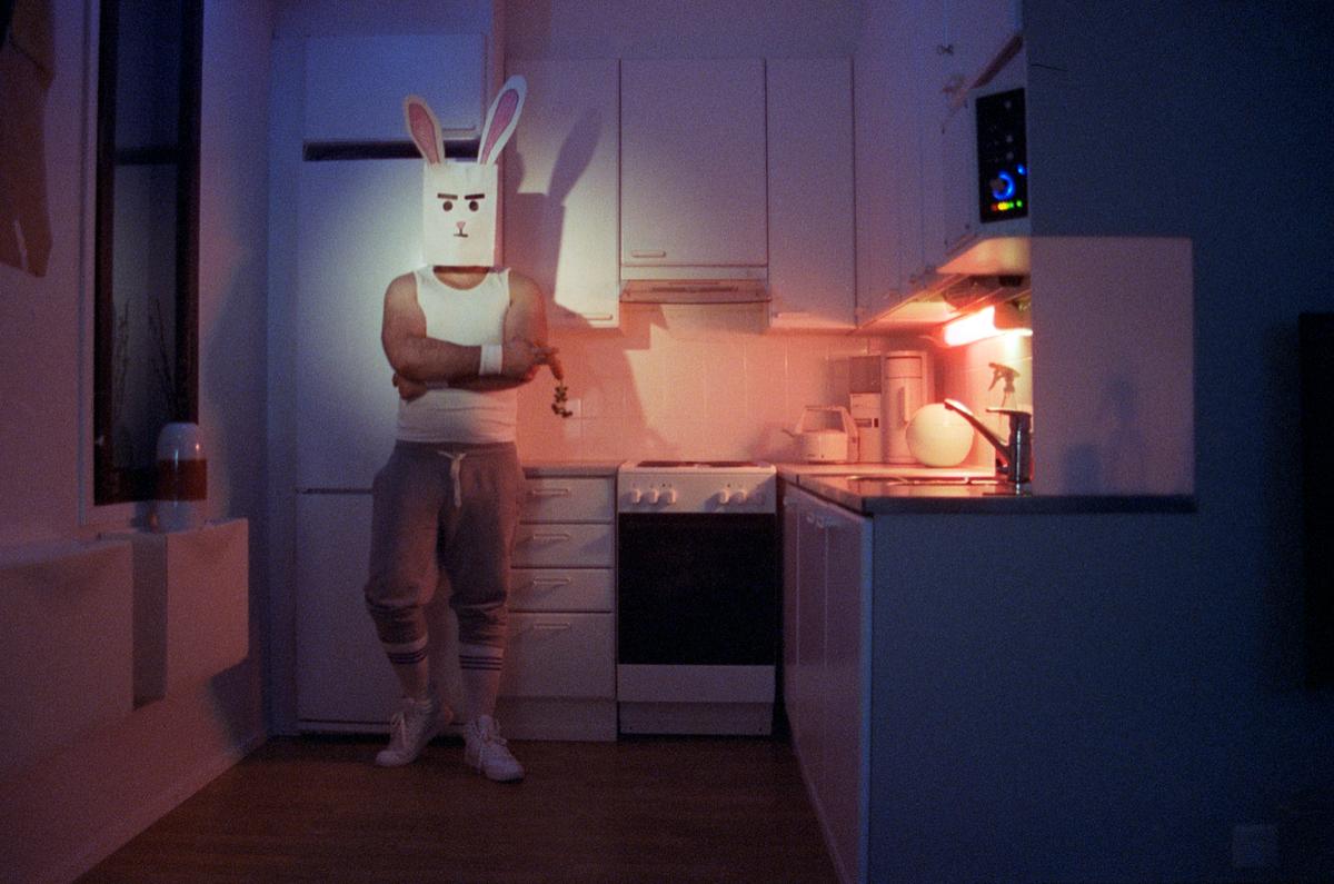 A man wearing a rabbit mask stands in a kitchen with his arms crossed.