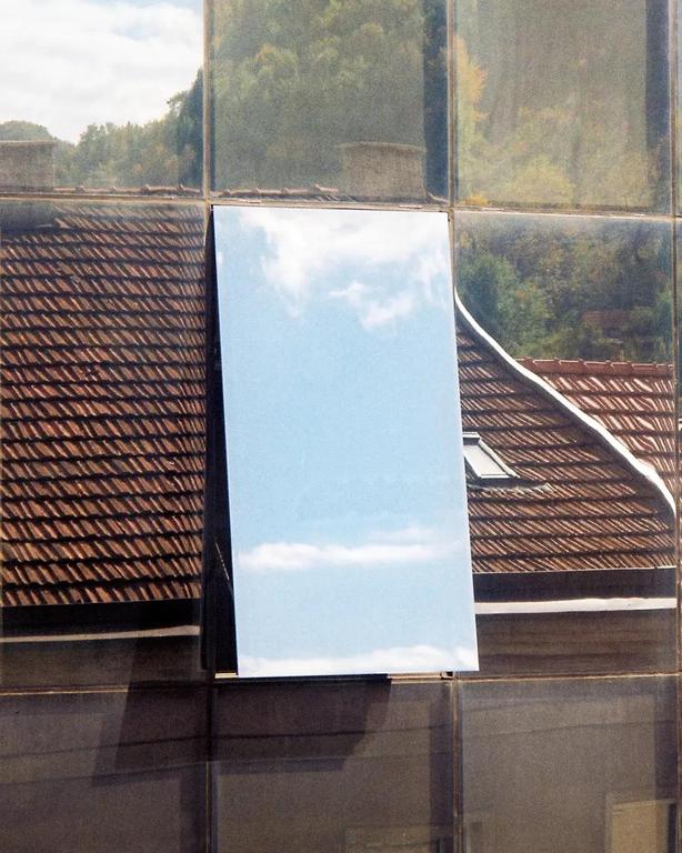 A mirror reflects the sky and clouds, surrounded by reflections of a tiled roof and trees.