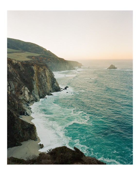 Waves crash against a rocky coastline with a sandy beach and a distant headland under a hazy sky.