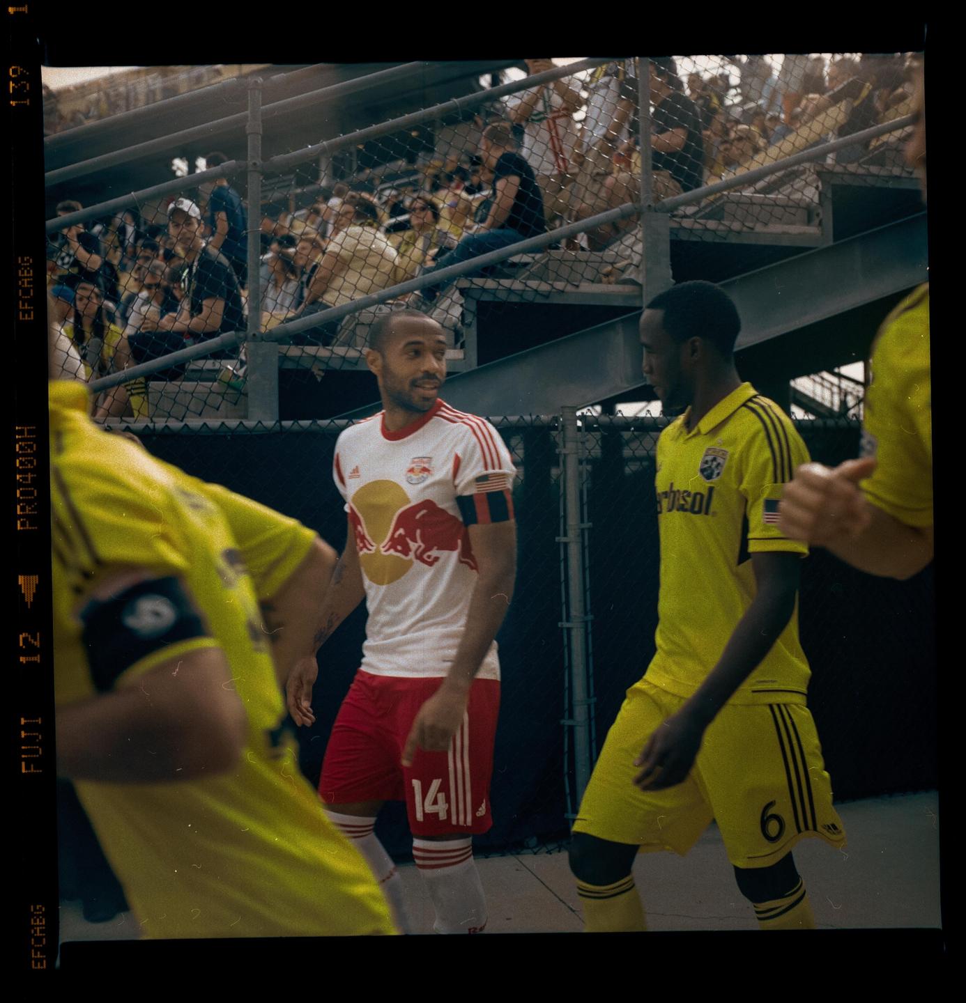 Three men in soccer uniforms are on a field with spectators in the background.