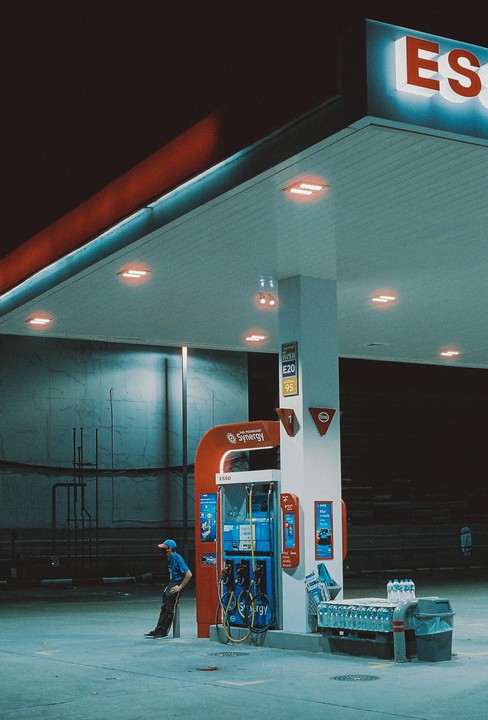 A man in a blue uniform leans against a pillar at a gas station at night.