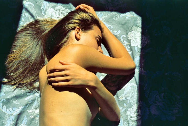 A woman with long hair lies on a patterned bedspread with her eyes closed, illuminated by a shaft of sunlight.