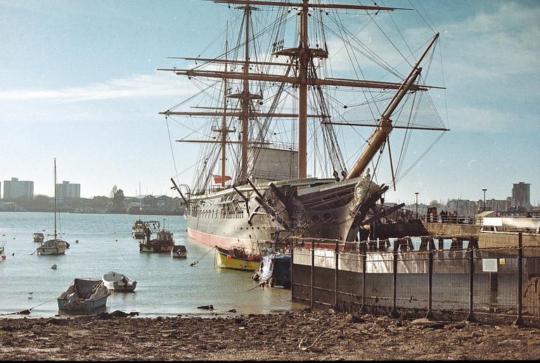 A large sailing ship is docked in a harbor with smaller boats in the water and buildings on the distant shore.