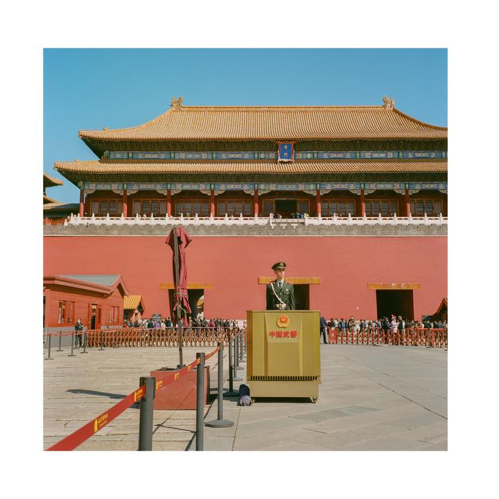 A uniformed man stands behind a golden podium in front of a red wall and a traditional Chinese building.